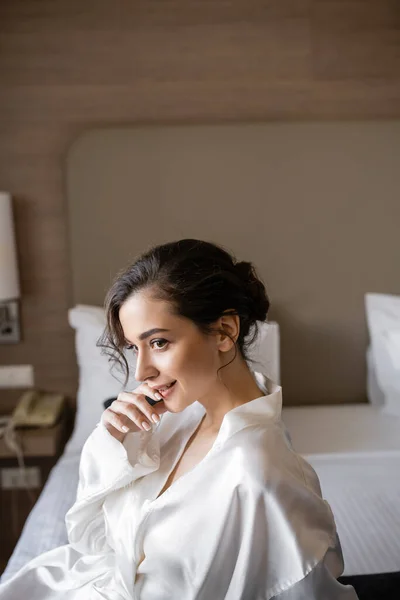 Happy young bride with brunette hair and elegant makeup sitting in white silk robe on comfortable bed and looking away in modern hotel suite on wedding day, special occasion — Stock Photo