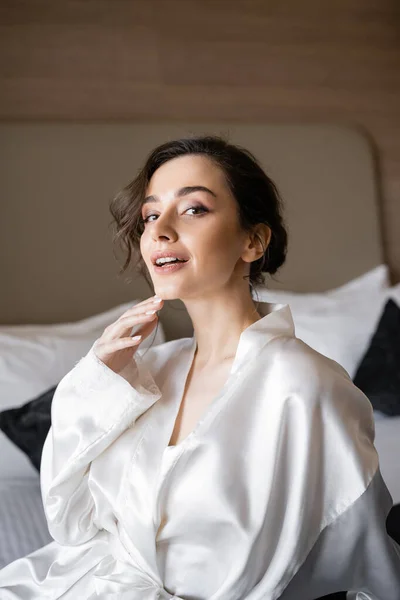 Anmutige junge Braut mit brünetten Haaren und Brautschminke, die das Kinn berührt und in einem weißen Seidenmantel auf einem bequemen Bett sitzt und am Hochzeitstag in die Kamera schaut — Stockfoto