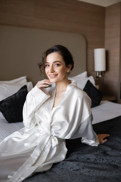 Pleased bride with brunette hair and bridal makeup touching chin and sitting in white silk robe on comfortable bed and looking at camera in hotel suite on wedding day — Stock Photo