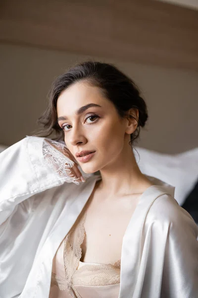 Enchanting woman with brunette hair and elegant bridal makeup posing with hand near face in white silk robe and looking at camera in hotel suite on wedding day, special occasion — Stock Photo