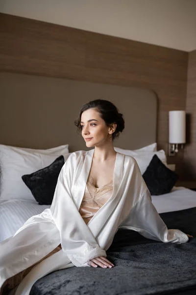 Charmante Braut mit brünetten Haaren und Brautschminke sitzt in weißer Seidenrobe auf bequemem Bett und schaut vor ihrer Hochzeit in der Hotelsuite weg, besonderer Anlass — Stockfoto