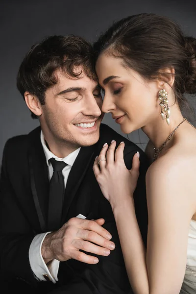 Portrait of bride with brunette hair, elegant jewelry and white dress hugging shoulder of happy groom in classic black suit with tie in modern hotel suite after wedding ceremony — Stock Photo