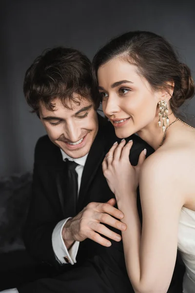 Portrait of bride with brunette hair, elegant jewelry and white dress hugging shoulder of happy groom in classic formal wear with tie in modern hotel suite after wedding ceremony — Stock Photo