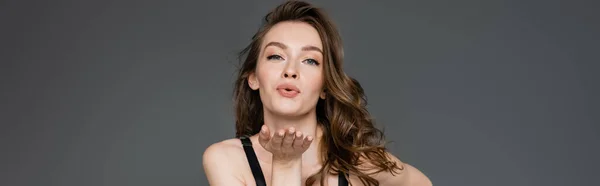 Pretty young model with natural makeup and brunette hair blowing air kiss while flirting and looking at camera while standing isolated on grey, banner — Stock Photo