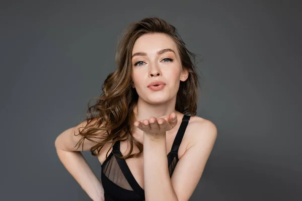 Seductive young brunette model with natural makeup and hairstyle wearing black bodysuit and looking at camera while blowing air kiss and flirting isolated on grey — Stock Photo