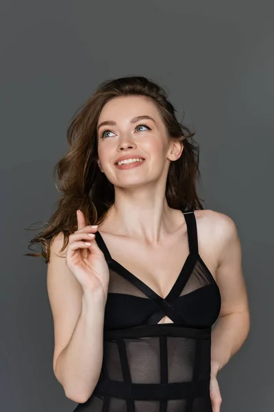 Портрет улыбающейся и сексуальной молодой женщины с натуральным макияжем, смотрящей в сторону и указывающей пальцем, позируя в черном и стильном телесном костюме, изолированном на сером — стоковое фото