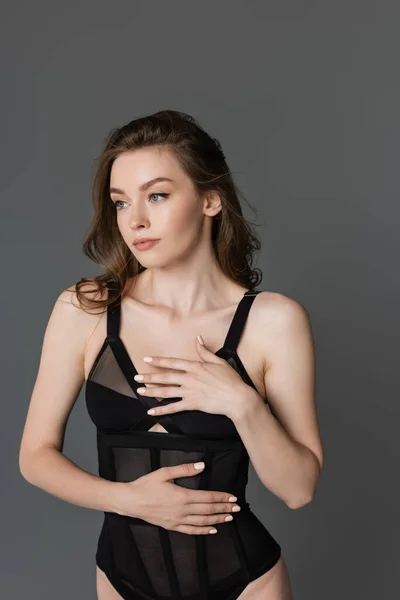 Portrait of sexy and brunette woman with natural makeup posing in black and trendy, form-fitting bodysuit and touching chest while looking away and standing isolated on grey — Stock Photo