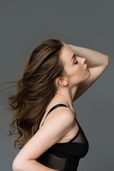 Side view of sexy and stylish young woman with natural makeup and slim body touching brunette hair while posing in black bodysuit and standing isolated on grey — Stock Photo