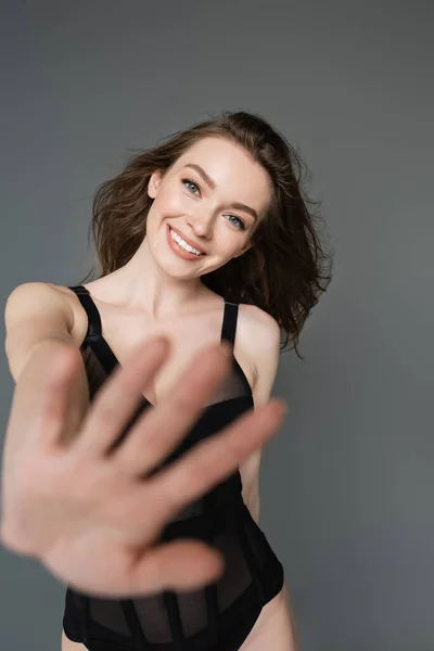 Portrait of smiling young brunette woman with natural makeup in trendy black form-fitting bodysuit outstretching hand and looking at camera while standing isolated on grey — Stock Photo