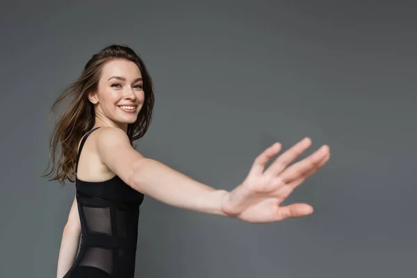 Positive and confident young brunette woman with natural makeup wearing stylish and sexy black and form-fitting bodysuit while looking at camera and standing isolated on grey, outstretched hand — Stock Photo