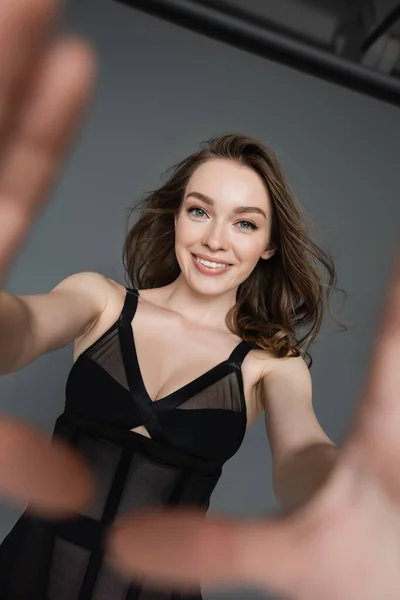 Portrait of cheerful and confident woman with natural makeup and slim body wearing sexy black bodysuit while looking and touching camera on grey background, blurred hands — Stock Photo