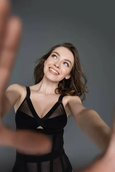Smiling and fashionable young brunette woman with natural makeup in sexy black bodysuit looking away while posing and standing isolated on grey, blurred hands — Stock Photo