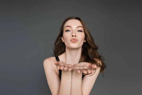 Pretty and young brunette model with hairstyle and natural makeup looking at camera while blowing air kiss while standing and posing isolated on grey,  flirt, chic — Stock Photo