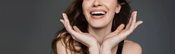 Cropped view of young and smiling brunette woman with everyday makeup holding hands near face while posing and standing with hands near face isolated on grey, banner — Stock Photo