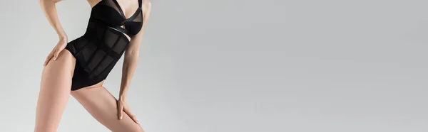 Vista cortada do modelo jovem sedutor no bodysuit preto elegante tocando quadril e perna enquanto em pé e posando isolado no cinza com espaço de cópia, banner — Fotografia de Stock