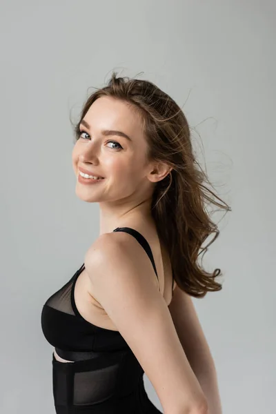 Portrait of positive and pretty young brunette woman with brunette hair and natural makeup looking at camera while posing in black bodysuit isolated on grey — Stock Photo