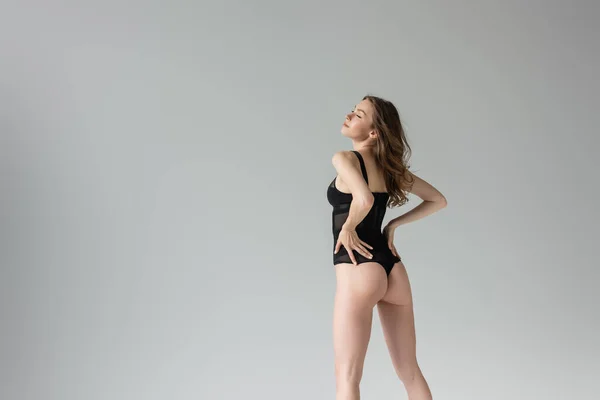 Full length of trendy and stylish young woman with closed eyes and brunette hair wearing sensual black bodysuit and boots while touching hips and standing isolated on grey — Stock Photo