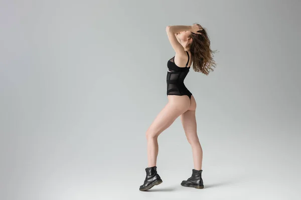 Longitud completa de la joven modelo de moda con el peinado tocando la cabeza mientras posa en el sensual traje negro y botas y de pie aislado en gris - foto de stock