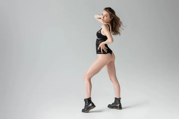 Full length of fashionable and sexy young woman with hairstyle posing in black bodysuit and boots and touching hip while looking at camera isolated on grey — Stock Photo