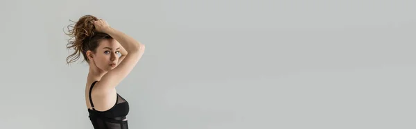 Sexy e confiante jovem mulher com maquiagem natural tocando cabelo morena e olhando para a câmera enquanto posando em corpo preto elegante isolado em cinza, banner — Fotografia de Stock