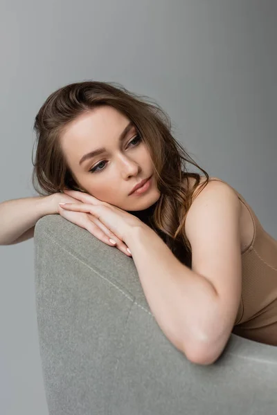 Portrait of young brunette woman with hairstyle and natural makeup wearing beige bodysuit and relaxing on comfortable grey armchair isolated on grey — Stock Photo