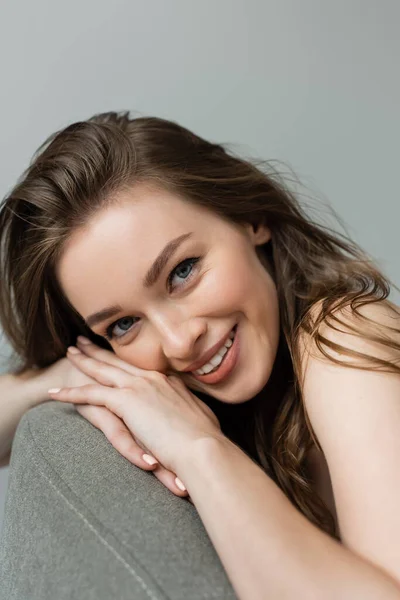 Retrato de una joven sonriente y guapa con maquillaje natural y cabello moreno mirando a la cámara mientras se relaja en un cómodo sillón gris aislado en gris - foto de stock