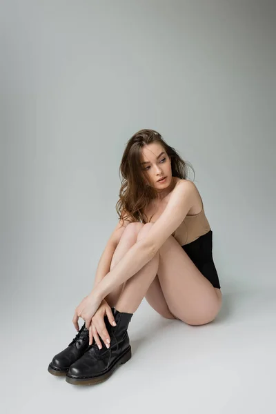 Full length of sexy and fashionable young woman in black boots, beige bodysuit and corset looking away while sitting and posing on grey background — Stock Photo