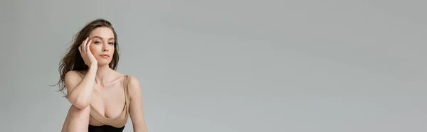 Giovane donna bruna alla moda con trucco quotidiano che tocca il viso mentre posa in body beige e corsetto nero e guarda la fotocamera isolata su grigio con spazio copia, banner — Foto stock