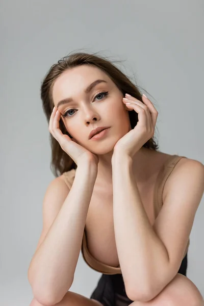 Portrait of young and seductive woman with brunette hair and natural makeup wearing bodysuit while touching face and looking at camera isolated on grey — Stock Photo