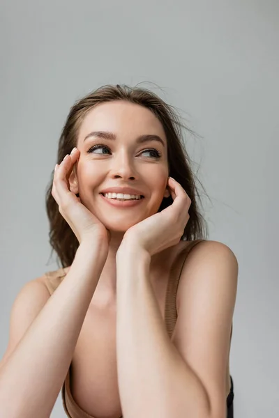 Portrait of cheerful and sexual young brunette woman with everyday makeup wearing bodysuit and looking away while posing isolated on grey, hands near face — Stock Photo