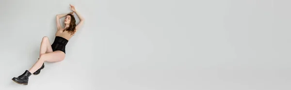 Modische und verführerische junge Frau in beigem Body, schwarzem Korsett und trendigen Stiefeln entspannt mit geschlossenen Augen auf grauem Hintergrund mit Kopierraum liegend, Banner — Stockfoto