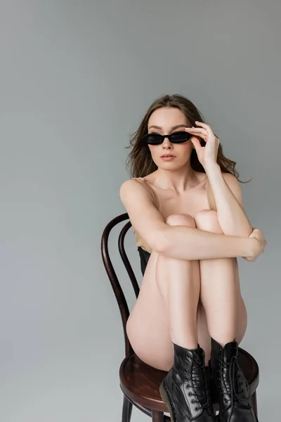 Модная молодая женщина с прической в телесном костюме и черных сапогах и трогательными стильными солнцезащитными очками, сидя на деревянном кресле, изолированном на сером — стоковое фото