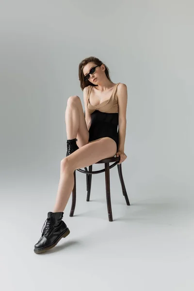 Full length of fashionable and young brunette model in sunglasses, black boots and beige bodysuit with sexy corset posing on wooden chair on grey background — Stock Photo