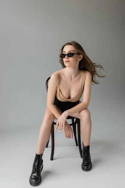 Full length of trendy young brunette model in sunglasses, beige bodysuit, black corset and boots posing while sitting on wooden chair on grey background — Stock Photo