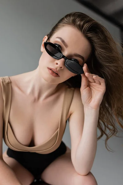 Porträt einer stilvollen und sexy brünetten Frau mit natürlichem Make-up, die Sonnenbrille berührt und in die Kamera schaut, während sie in beigem Body und Korsett auf grauem Hintergrund posiert — Stockfoto