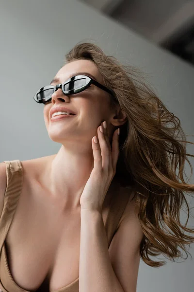 Low angle view of happy and seductive brunette model in stylish sunglasses smiling and touching face while posing in beige bodysuit on grey background — Stock Photo