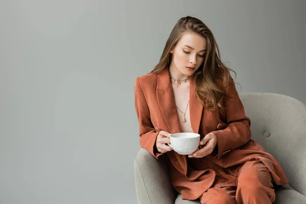 Mulher elegante jovem em jaqueta de terracota e calças usando colares e segurando xícara de cappuccino enquanto sentado em poltrona confortável isolado em cinza — Fotografia de Stock