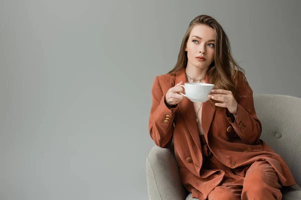 Trendige junge und brünette Frau im Terrakottaanzug hält eine Tasse Cappuccino in der Hand und schaut weg, während sie auf einem modernen Sessel sitzt, isoliert auf grau mit Kopierraum — Stockfoto