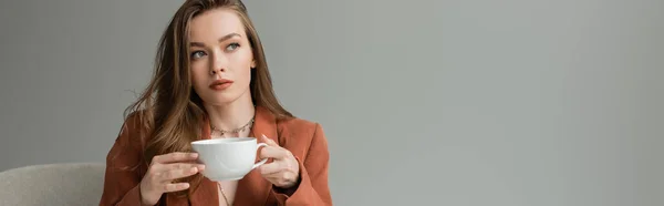 Elegante und stylische brünette Frau in Terrakottamaske und Halsketten, die eine Tasse Cappuccino in der Hand hält und wegschaut, während sie auf einem Sessel sitzt, isoliert auf grau, Banner — Stockfoto