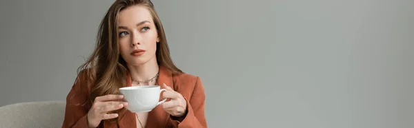 Mulher morena elegante e elegante em jaqueta de terracota e colares segurando xícara de cappuccino e olhando para longe enquanto sentada em poltrona isolada em cinza, banner — Fotografia de Stock