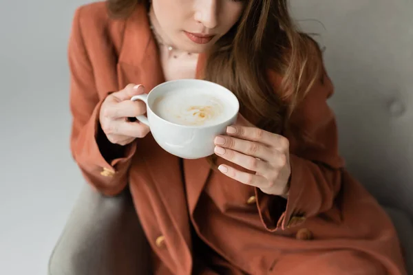 Vue recadrée d'une élégante jeune femme brune en blazer en terre cuite tenant une tasse de cappuccino assise sur un fauteuil flou et confortable isolé sur du gris — Photo de stock