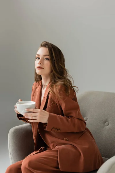 Modische junge Frau in Terrakotta-Anzug und Halskette blickt in die Kamera, während sie eine Tasse Cappuccino in der Hand hält und auf einem modernen Sessel sitzt, der isoliert auf grau ist — Stockfoto
