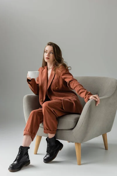 Брюнетка молода жінка з довгим волоссям і намистом в теракоті і модний костюм з блейзером і штанами і тримає чашку кави, сидячи в зручному кріслі на сірому фоні в студії — стокове фото