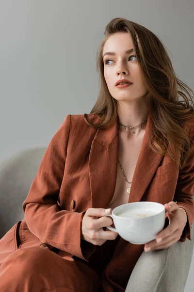 Brunette young woman with long hair and necklace wearing terracotta and trendy suit with blazer and pants and holding cup of coffee with foam while sitting in comfortable armchair on grey background — Stock Photo