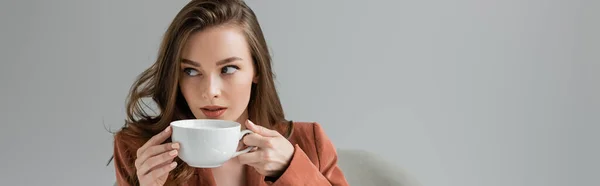 Brunette young woman with long hair and necklace wearing trendy suit with blazer and drinking coffee while looking away on grey background in studio, sophisticated lady, coffee break, banner — Stock Photo