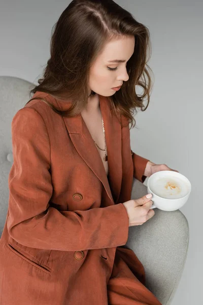 Brünette junge Frau mit langen Haaren und Halskette im schicken Anzug und mit einer Tasse Cappuccino im bequemen Sessel auf grauem Hintergrund im Studio sitzend, Kaffeepause, entspannt — Stockfoto