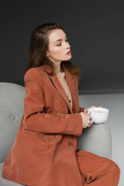 Brunette young woman in golden necklace wearing brown and trendy suit and holding cup of cappuccino while sitting in comfortable armchair on grey background in studio, coffee break, look away — Stock Photo