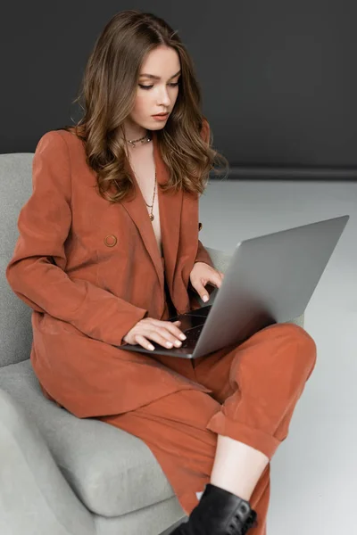 Morena joven con el pelo largo y collar con traje de moda con chaqueta y pantalones usando el ordenador portátil mientras está sentado en un cómodo sillón sobre fondo gris, freelancer, trabajo remoto - foto de stock