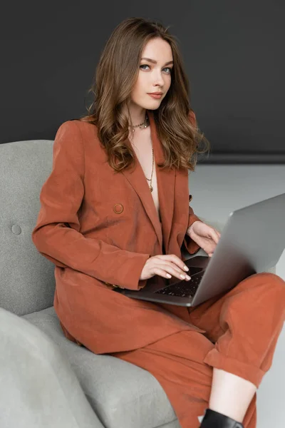 Mujer joven con el pelo largo y collar con traje de moda de terracota con chaqueta usando el ordenador portátil mientras está sentado en cómodo sillón sobre fondo gris, freelancer, trabajo remoto, mira a la cámara - foto de stock