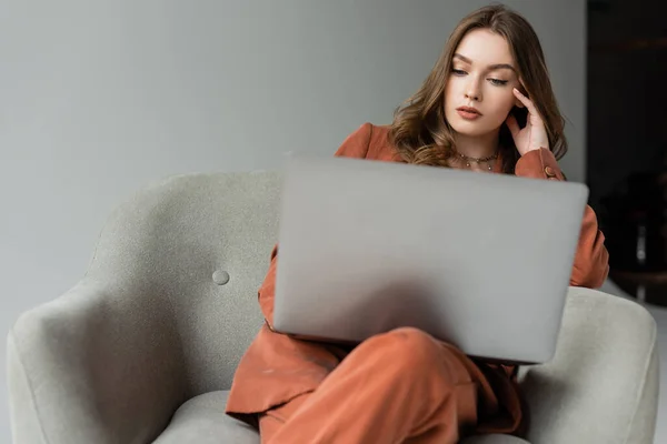 Brunette young woman with long hair and necklace wearing terracotta trendy suit with blazer and pants and using laptop while sitting in comfortable armchair on grey background, freelancer, remote work — Stock Photo